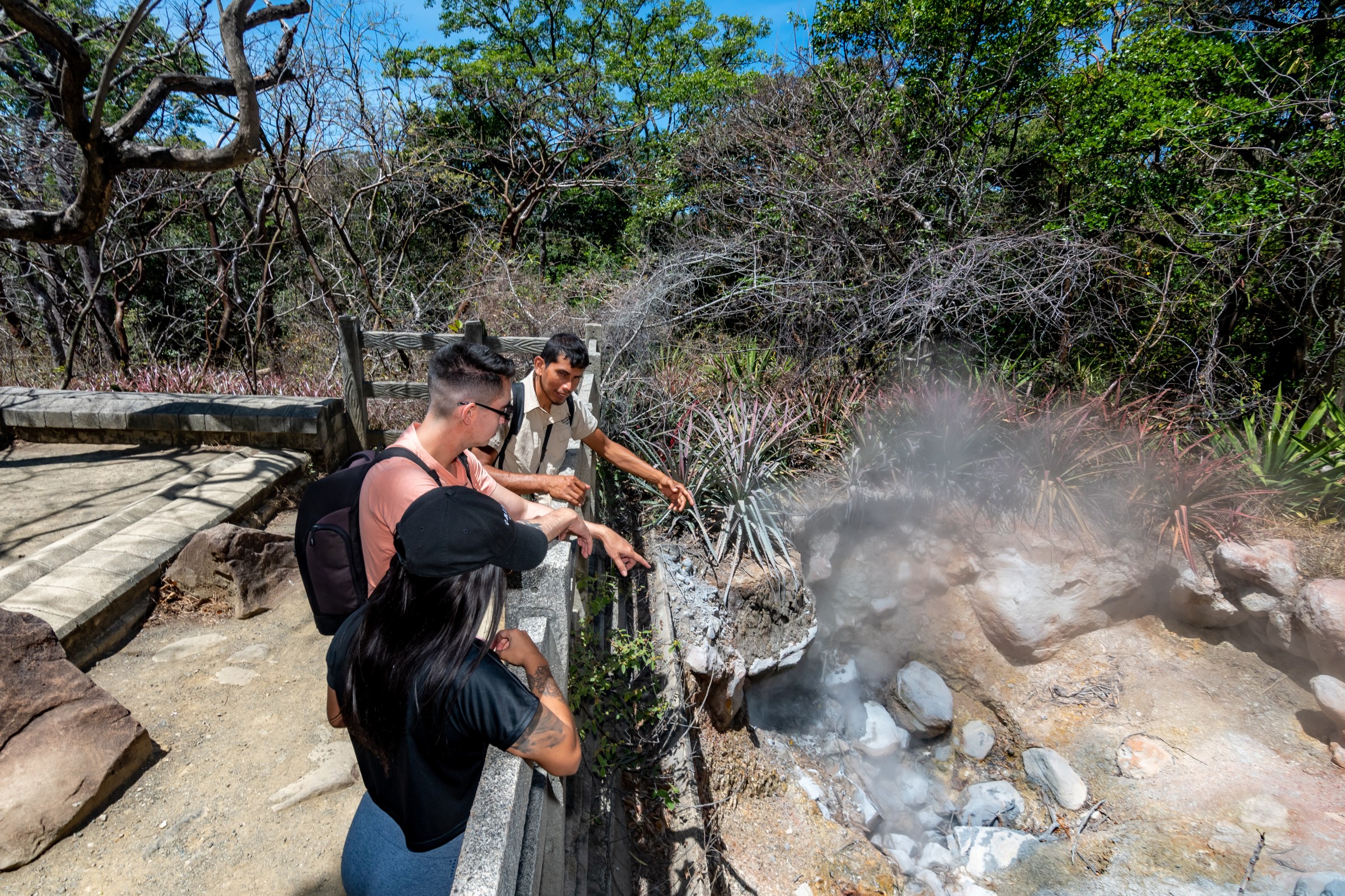 People observing steaming geothermal area with guide in tropical setting.