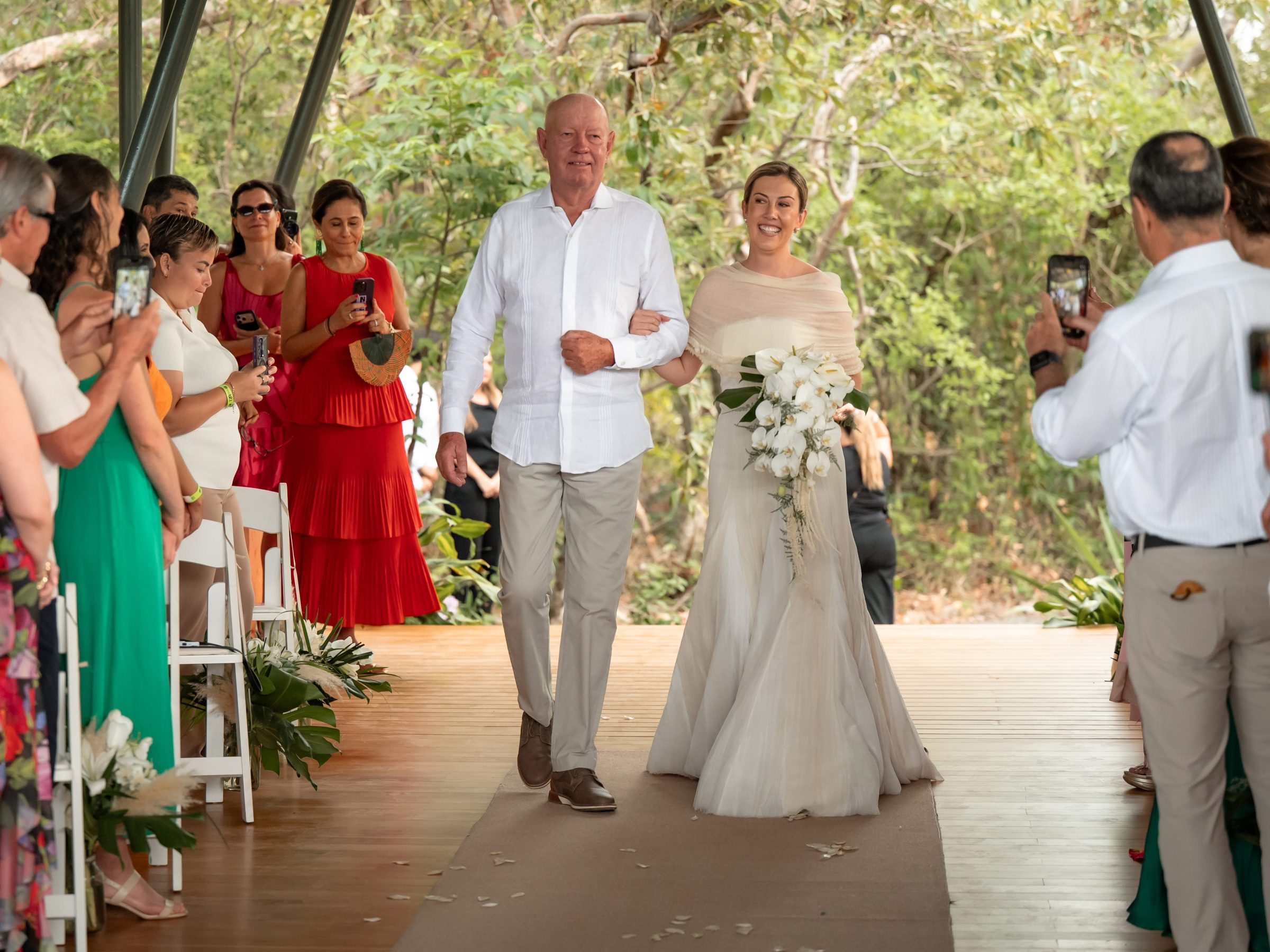 Man escorting a smiling bride down an outdoor aisle with guests taking photos.