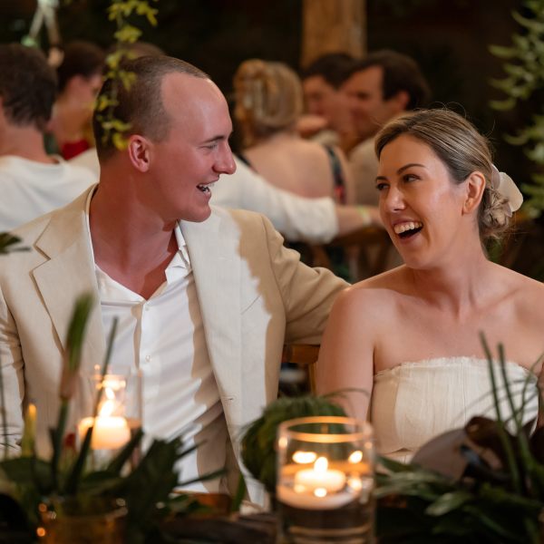 Smiling couple sitting at a wedding reception table with candles and greenery.
