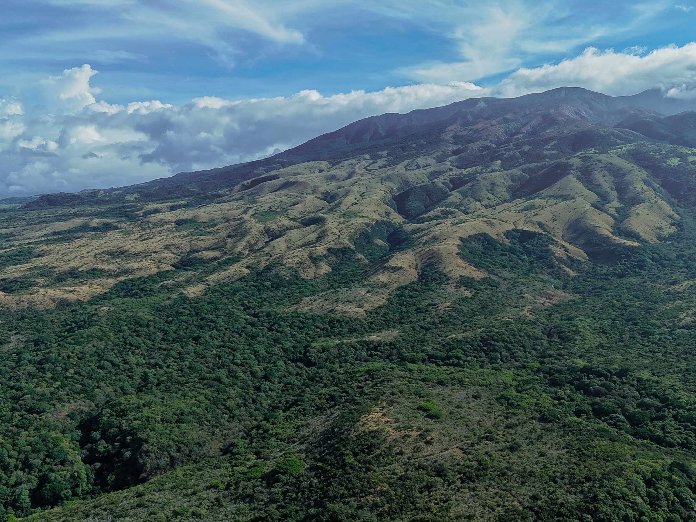 Aerial view of a mountain range with forests under a cloudy blue sky.