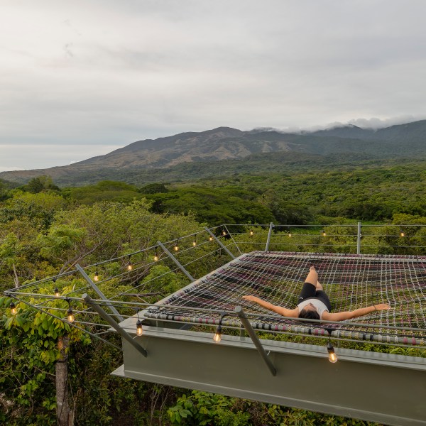 Person relaxing on a suspended net above lush greenery with mountains in the background.