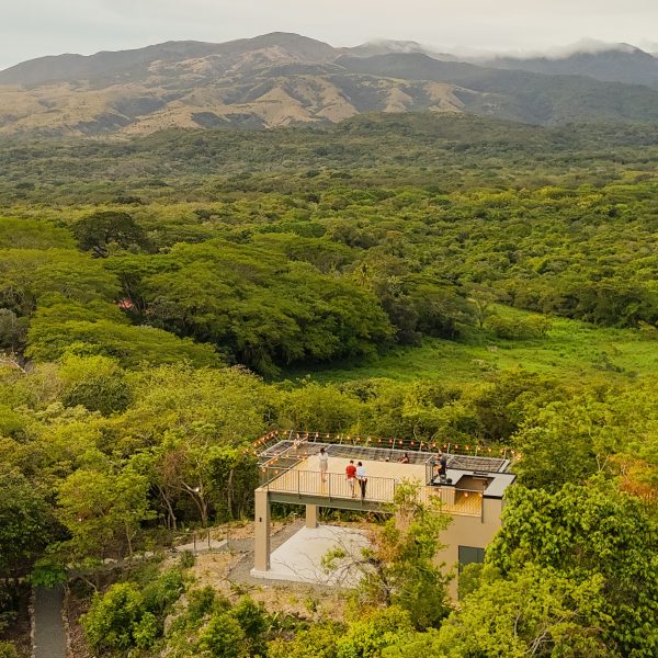 Aerial view of a house surrounded by dense forest with mountains in the background.