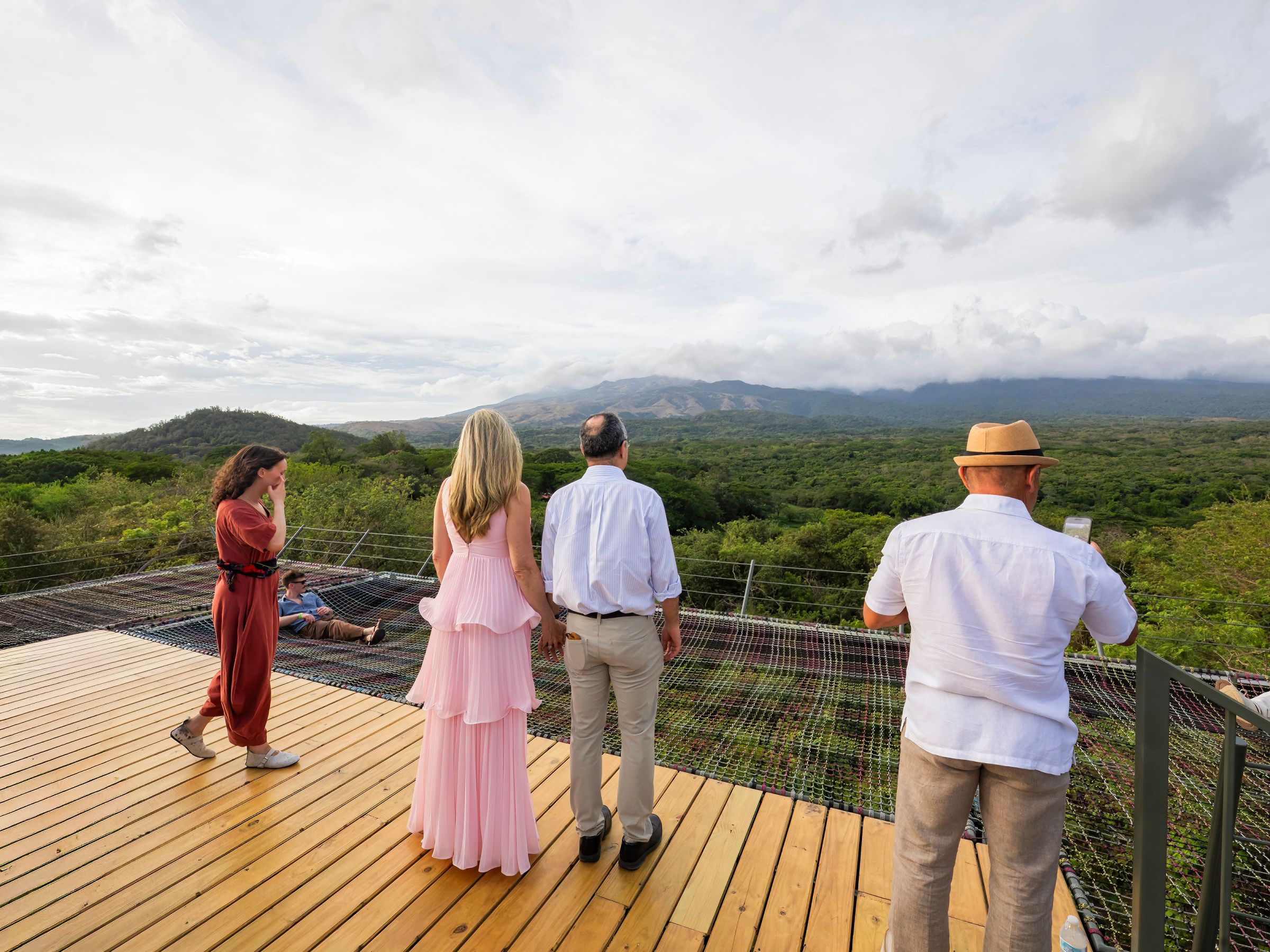 People on a wooden deck overlooking a green forest and distant mountains under a cloudy sky.