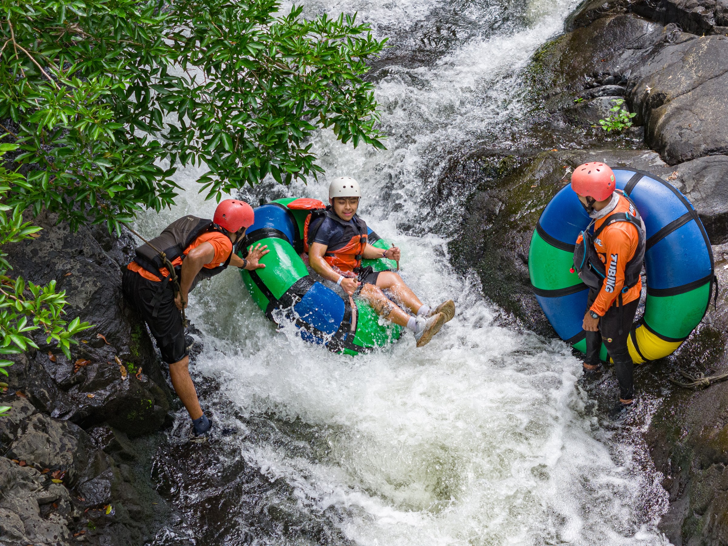 Three people river tubing on a rocky stream, wearing helmets and vests.