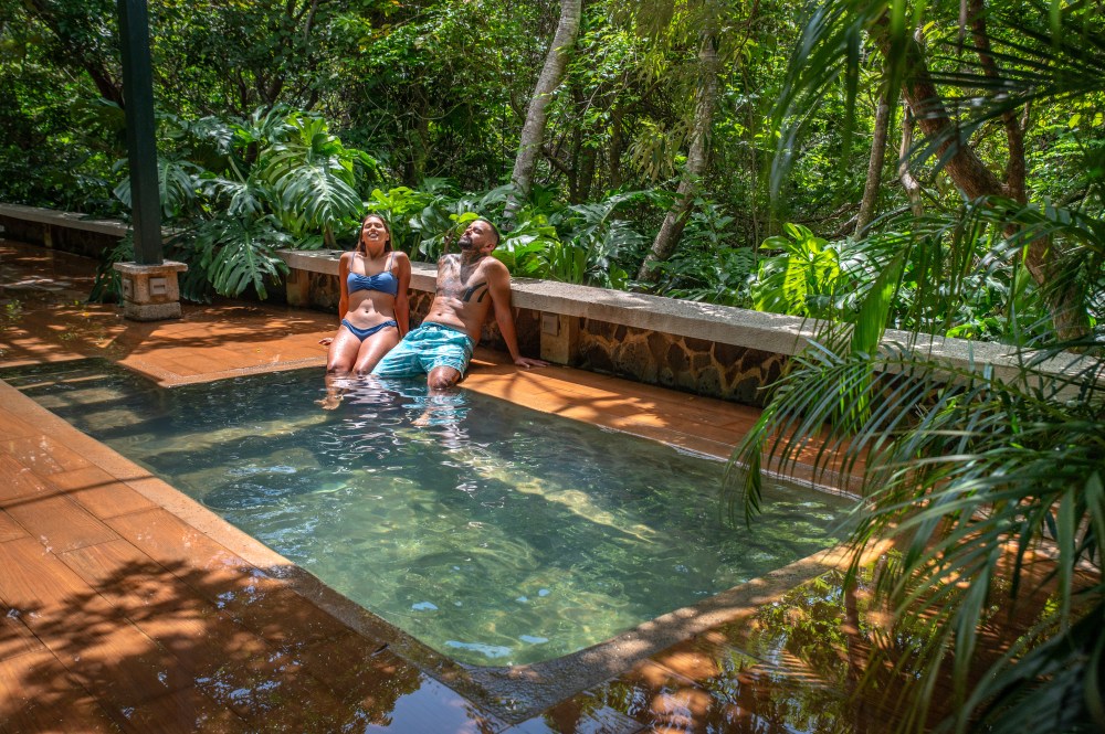 Two people relaxing in a small pool surrounded by lush greenery.
