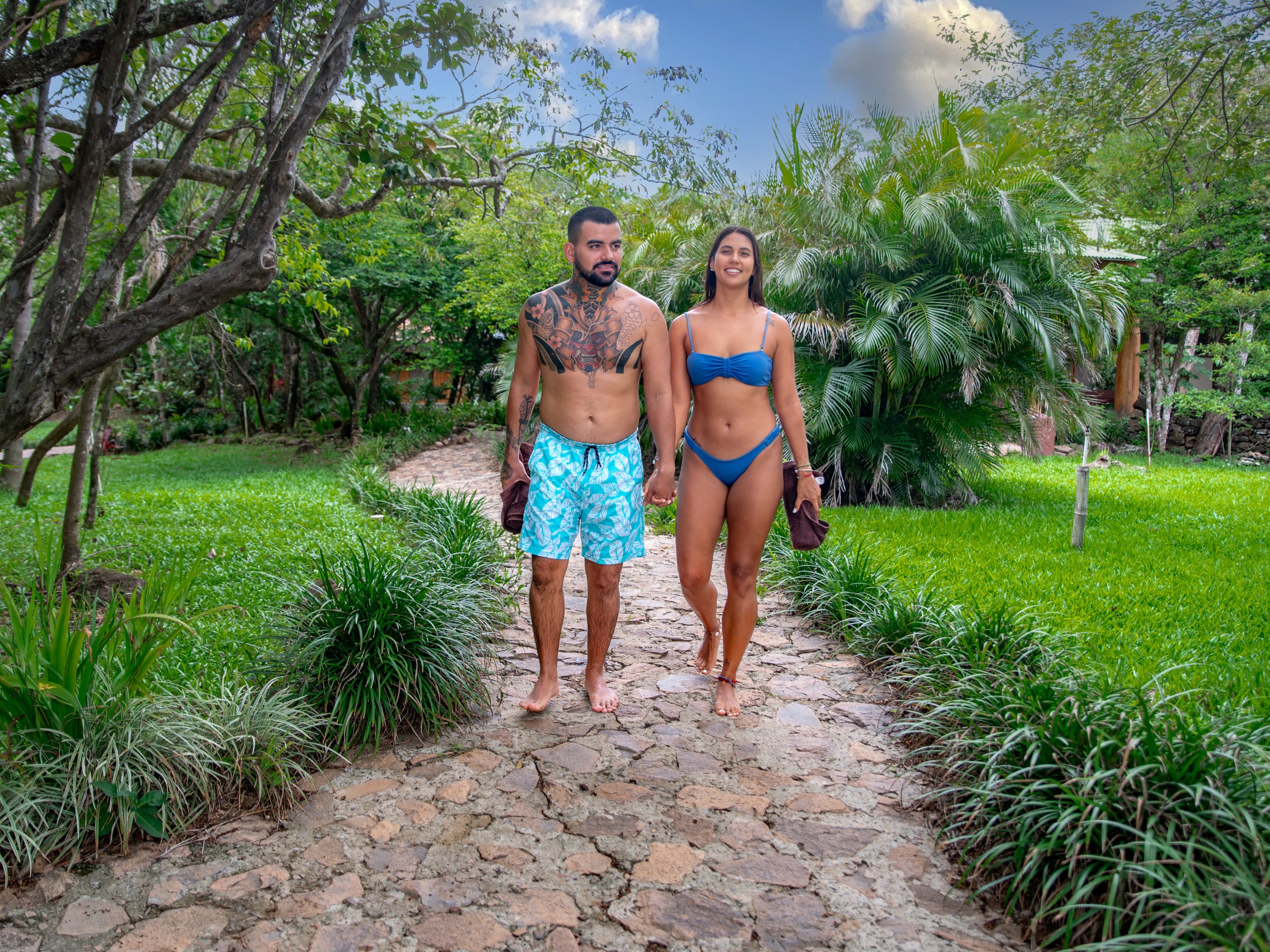 Man and woman in swimwear walking on a stone path in a lush garden.