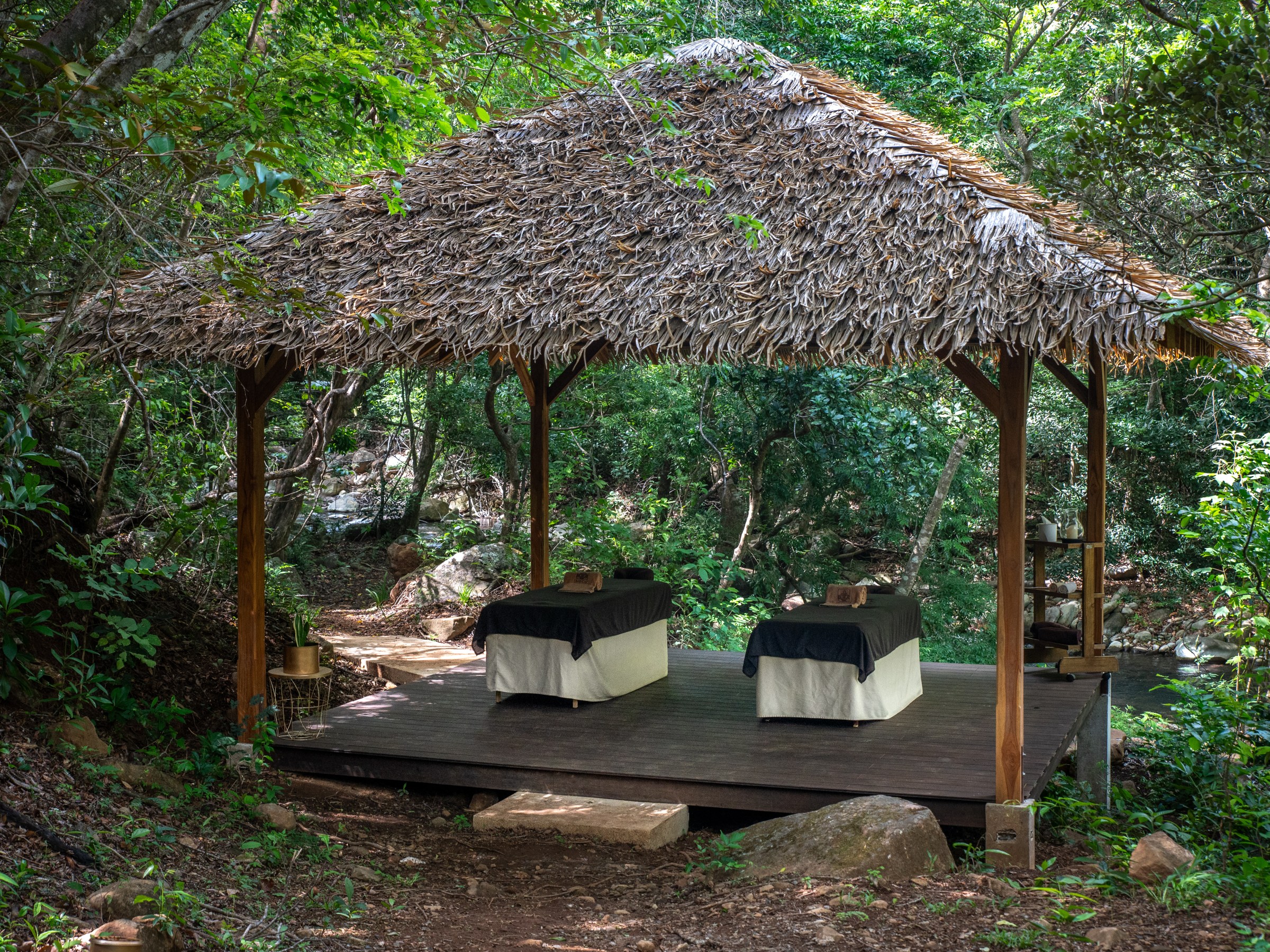 Outdoor spa with two tables under a thatched roof in a forest setting.