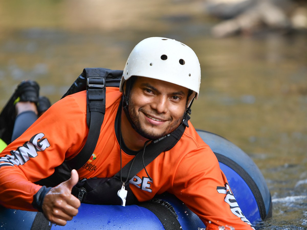 Person in orange gear and helmet smiles while lying on a blue raft in a river.