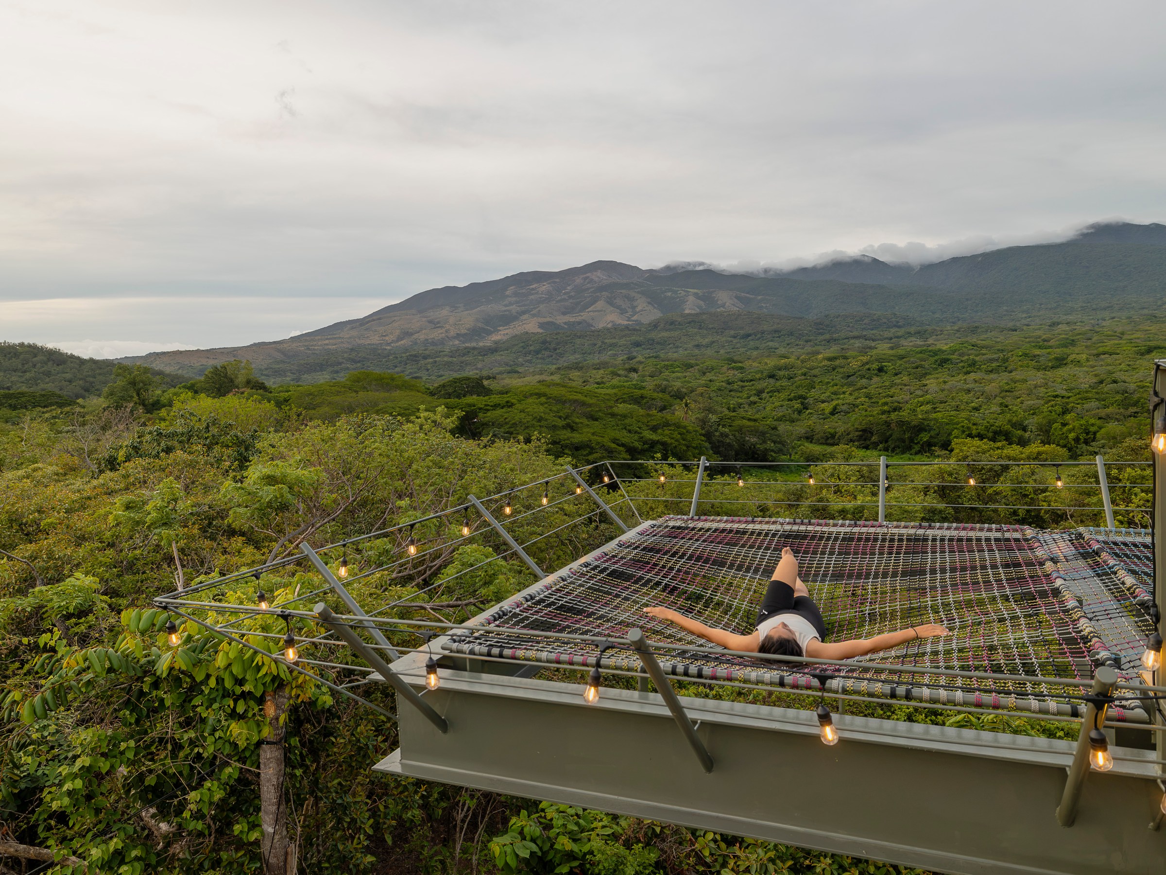 Person laying on a hammock above lush greenery with mountains in the background.