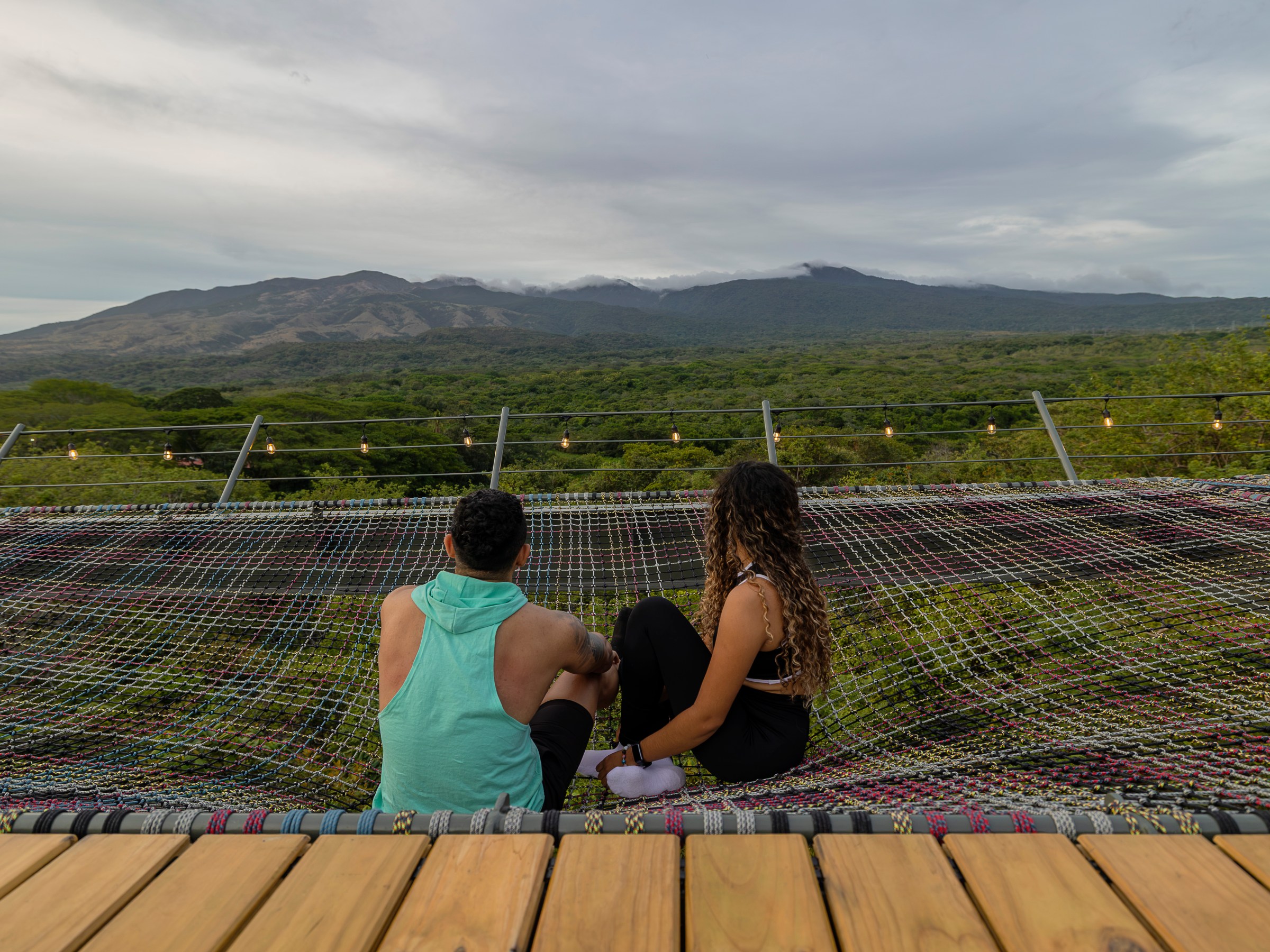 Two people sit on a netting, overlooking a scenic mountain view.