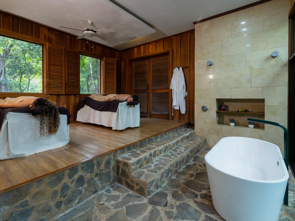Two people on massage tables in a serene spa room with bathtub and large windows.