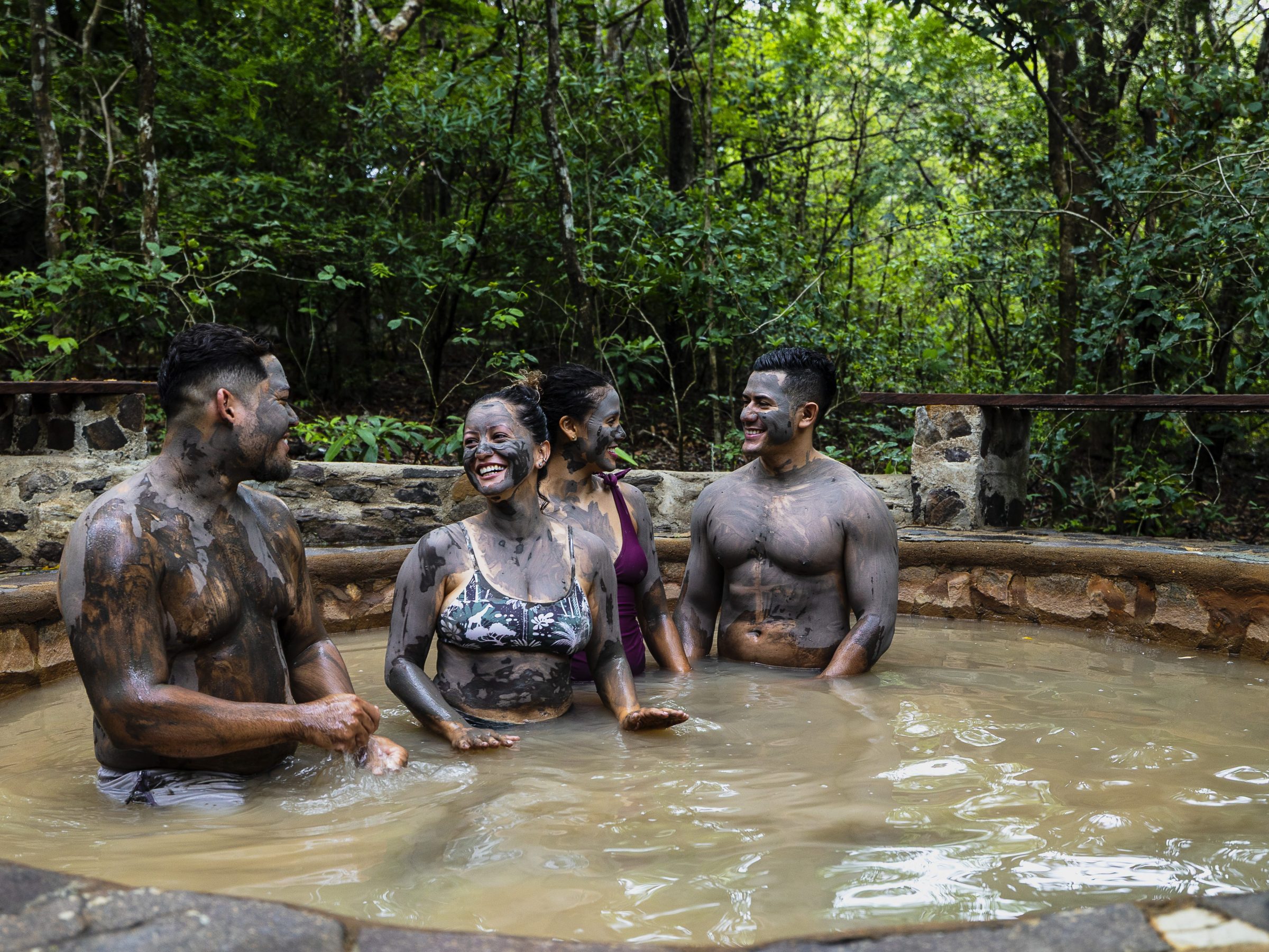 Four people covered in mud laughing in a forest hot spring.